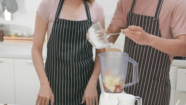 Happy Asian beautiful young family couple husband and wife making fresh apple smoothie in kitchen together at home. The man and woman pour ice into a juice maker blender. Healthy lifestyle concept