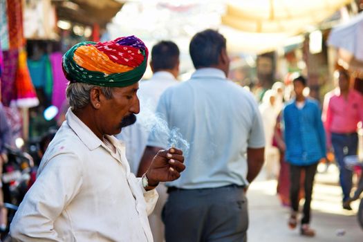 Pushkar, India - NOVEMBER 10, 2016: An old Rajasthani man in traditional ethnic wear such as colorful turban and typical white shirt smoking cigarette or mini-cigar filled with tobacco