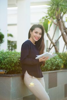 Portrait of smiling female student reading book while standing outdoor on terrace of campus cafe in sunny day. Education, lifestyle and people concept.