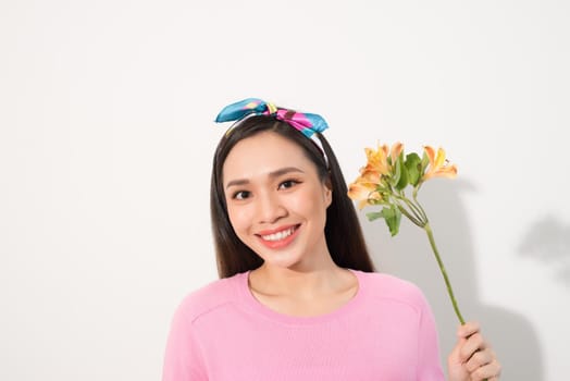 Close up portrait of adorable, beautiful, cute woman in casual clothes with beaming smile and long blond hair, holding three flowers in hand near cheek, standing over white background