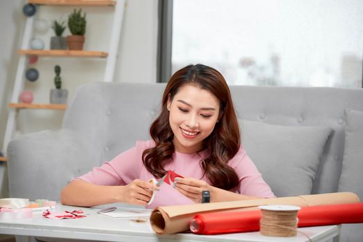 Smiling girl cutting valentine heart out of red paper with scissors in living room