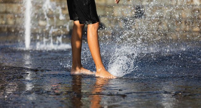 Happy children playing in a water fountain in a hot day. Close-up of children legs in a fountain