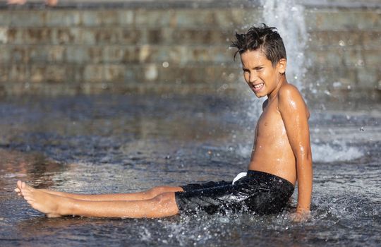 11 years old happy boy playing in a water fountain and enjoying the cool streams of water in a hot day. Hot summer.