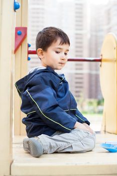 Serious cute boy sits on playground with houses on background