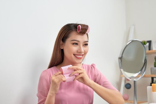 Head and shoulders portrait of beautiful Asian woman wearing hair curlers looking in mirror with wide smile, home interior on background