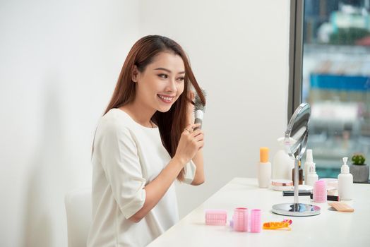 Getting rid of tangles. Beautiful young woman looking at her reflection in mirrorand brushing her long hair while sitting at the dressing table