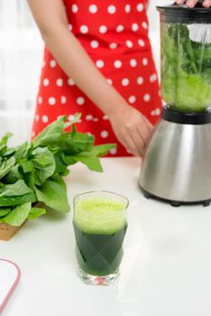 Young smiling woman making smoothie with fresh greens in the blender in kitchen at home. Healthy vegetarian smoothie for weight loss and detox