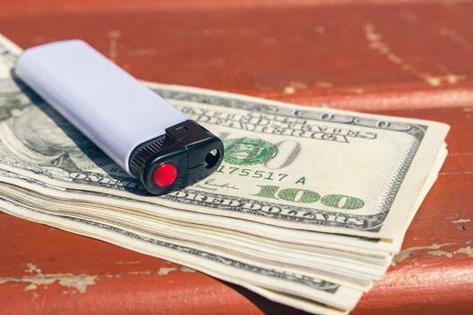 A pocket lighter rests on a stack of one hundred dollar bills on an outdoor bench