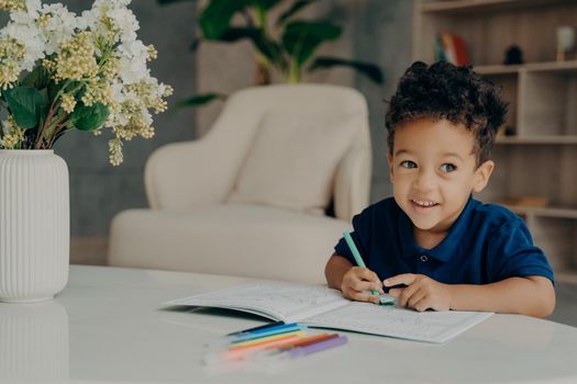 Happy curly mixed race boy coloring animals in coloring book with felt pens, looking aside and smiling while sitting in front of table in comfy living room at home. Children leisure time activities