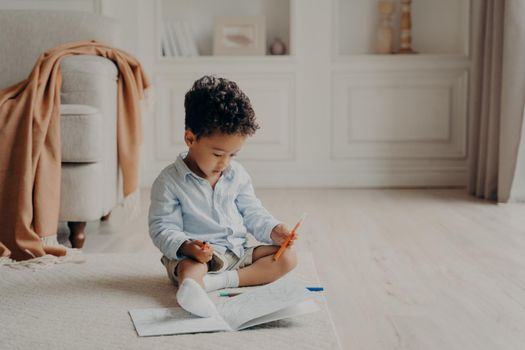 Small cute curly mulatto boy sitting on floor in living room in front of colouring book with felt tip pen in hand, child deciding which superhero to colour while spending leisure time at home