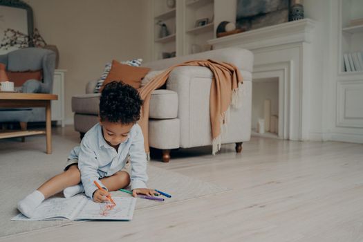 Little mixed race kid boy drawing in coloring book with lots of different felt tip pen while sitting alone on floor in middle of living room at home. Children leisure and developing activities