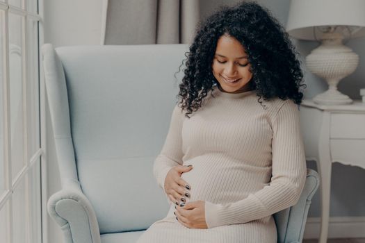 Happy pregnancy. Pretty mixed race woman with curly hair in white casual dress holding her belly in anticipation of future baby, thinking about maternity and how to be caring and loving mother