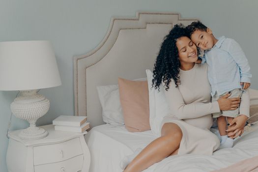 My little boy. Happy pregnant afro american woman in casual white dress with curly hair sitting on bed with her sweet little son and showing strong feelings of love and care. Maternity and pregnancy