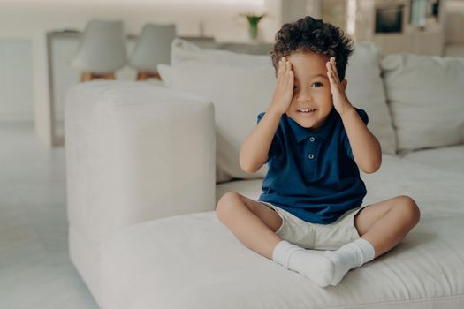 Adorable curly afro american boy in casual wear sitting on comfy big white couch and ready to play, stopping counting and uncovering his eyes, about to go searching in game of hide and seek