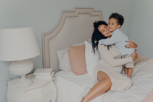 Happy family at home. Smiling pregnant mixed race woman mother in dress with curly hair hugging her beloved sweet little son while sitting on big soft bed. Mom and little child spending time together