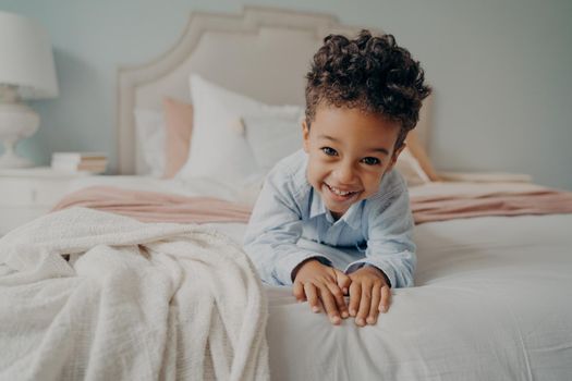 Portrait of happy curly mulatto boy smiling at camera while laying on cozy bed, relaxing after fun playing session at home, adorable preschool child enjoying indoor activities and leisure time