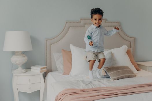 Happy little energetic afro american kid boy jumping alone on his parents bed while holding delicious lollipop, eating sweets, causing mess while no one is home, cute child having fun at home