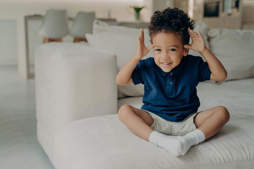 Full length shot of happy playful curly mulatto child holding his hands up, making funny face expression while sitting alone on couch, spending carefree and leisure time at home. Childhood concept