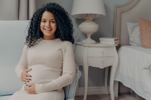 Portrait of young positive pregnant woman sitting in bedroom and looking at camera with bright smile, holding her belly and enjoying pregnancy time while relaxing on armchair at home