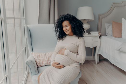 Charming young mixed race pregnant woman, future mother looking out of big window and dreaming about maternity while sitting sideways on armchair in bedroom, holding belly with both hands and smiling