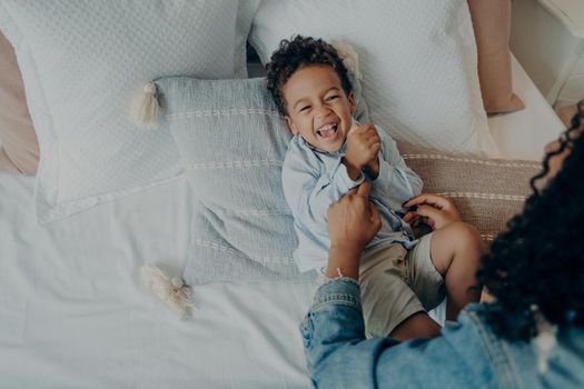 Sweet little boy kid laughing loudly and smiling while his loving mom tickling him on bed with lot of soft pillows, mother telling really funny story to son while spending happy time together at home