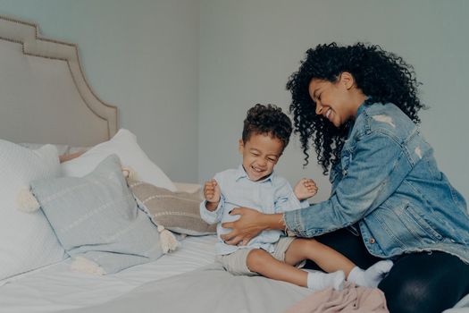 Joyful afro american mother with bright smile and curly hair tickling her cute little son toddler while spending happy time with each other in bedroom, loving mom and child having fun, playing indoor