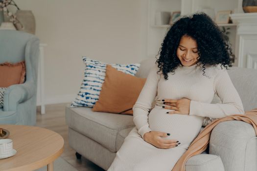 Happy young pregnant afro american lady in white dress with long curly hair looking at her belly with gentle smile, taking little break from housework by resting on couch and drinking cup of green tea