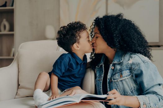 Beautiful afro american family mother with little son kissing each other and expressing love while spending time together at home, reading book while sitting on soft white couch in living room