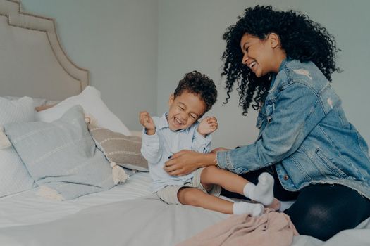 Happy mixed race family at home, carefree young woman mom in denim jacket and jeans with curly hair playing with her sweet child little boy, tickling his tummy while spending time together in bedroom