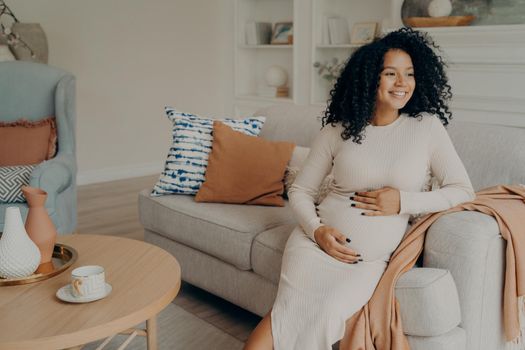 Happy pregnant afro american woman in casual dress with curly hair looking at window while sitting on couch in living room, checking out weather and thinking about going outside for walk