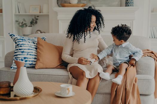 Lovely pregnant mixed race woman mother explaining to curious son about his future brother or sister, cute little boy touch belly of his mom and talking with baby while sitting on couch in living room