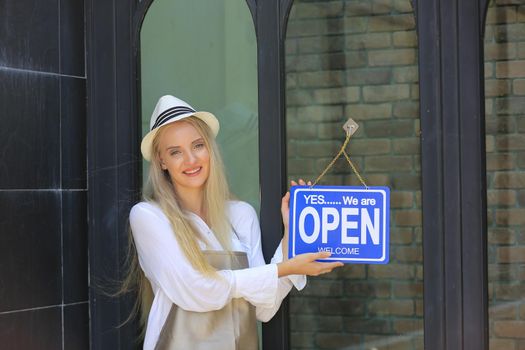 Beautiful blonde hair girl standing by open shop sign plate at door with confidence in front of coffee shop.