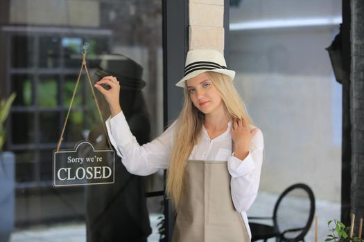 Beautiful blonde hair girl standing by open shop sign plate at door with confidence in front of coffee shop.