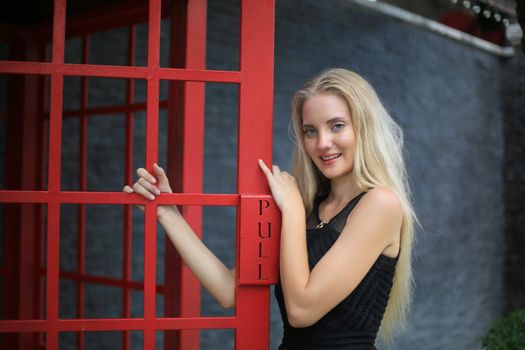 Portrait of Beautiful blonde hair girl on black dress standing in red phone booth against black wall as portrait fashion pose outdoor.