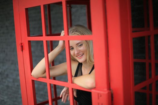 Portrait of Beautiful blonde hair girl on black dress standing in red phone booth against black wall as portrait fashion pose outdoor.