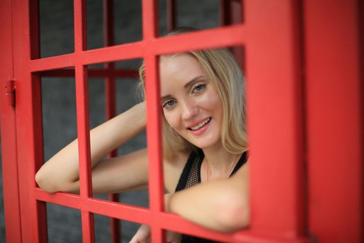 Portrait of Beautiful blonde hair girl on black dress standing in red phone booth against black wall as portrait fashion pose outdoor.