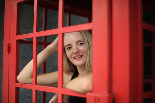 Portrait of Beautiful blonde hair girl on black dress standing in red phone booth against black wall as portrait fashion pose outdoor.