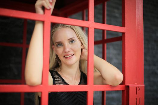 Portrait of Beautiful blonde hair girl on black dress standing in red phone booth against black wall as portrait fashion pose outdoor.