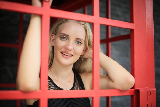 Portrait of Beautiful blonde hair girl on black dress standing in red phone booth against black wall as portrait fashion pose outdoor.