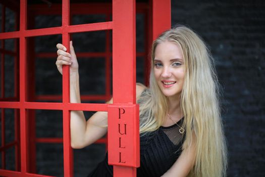 Portrait of Beautiful blonde hair girl on black dress standing in red phone booth against black wall as portrait fashion pose outdoor.