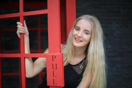 Portrait of Beautiful blonde hair girl on black dress standing in red phone booth against black wall as portrait fashion pose outdoor.