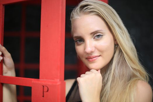 Portrait of Beautiful blonde hair girl on black dress standing in red phone booth against black wall as portrait fashion pose outdoor.