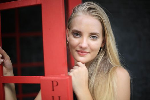 Portrait of Beautiful blonde hair girl on black dress standing in red phone booth against black wall as portrait fashion pose outdoor.