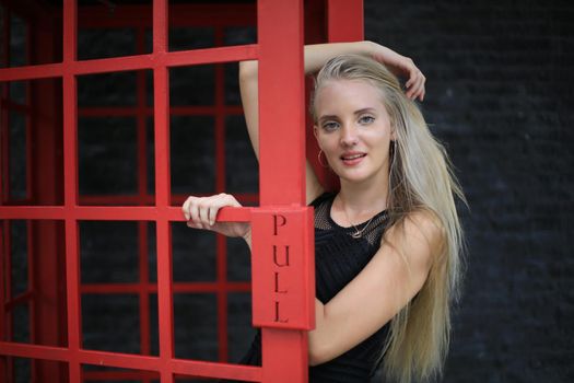 Portrait of Beautiful blonde hair girl on black dress standing in red phone booth against black wall as portrait fashion pose outdoor.