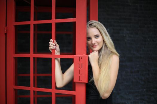 Portrait of Beautiful blonde hair girl on black dress standing in red phone booth against black wall as portrait fashion pose outdoor.