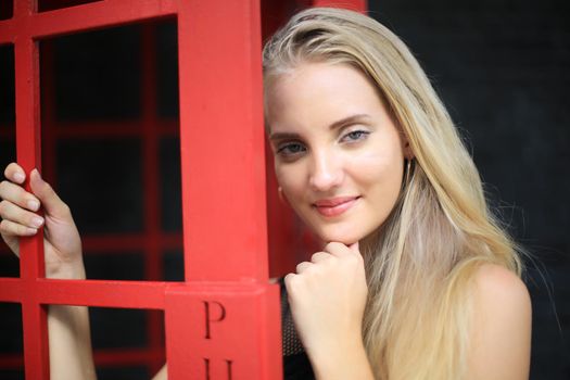 Portrait of Beautiful blonde hair girl on black dress standing in red phone booth against black wall as portrait fashion pose outdoor.