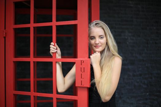 Portrait of Beautiful blonde hair girl on black dress standing in red phone booth against black wall as portrait fashion pose outdoor.