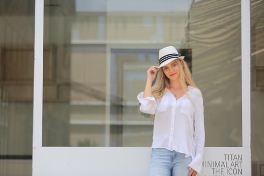 Beautiful blonde hair girl with white hat standing against wall glass as fashion pose outdoor.