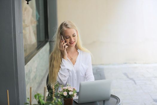 Beautiful blonde hair girl speaking on mobile phone with laptop on table outside coffee shop.