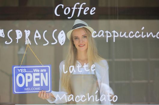Beautiful blonde hair girl standing by open shop sign plate at door with confidence in front of coffee shop.